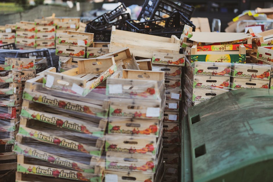 A large outdoor scene featuring multiple stacks of cardboard fruit boxes, most labeled with the text 'fresh fruits' and decorated with images of tomatoes and other produce, arranged on a paved surface. Some boxes are empty with visible wooden slats and handles, while others are flattened or partially collapsed. In the background, there are black plastic crates piled haphazardly, and a green plastic container is partially visible at the bottom right corner. The lighting appears natural, with even illumination across the scene, highlighting the textures of the cardboard, plastic, and pavement. The arrangement suggests a collection of discarded packaging material or surplus stock, a common scenario in waste management contexts where bulk fruit packaging is being sorted for disposal or recycling. This scene exemplifies typical outdoor clutter associated with rubbish clearance services, such as those provided by independent waste disposal specialists like Rubbish Clearances within Colliers Wood, addressing non-local authority waste handling needs.