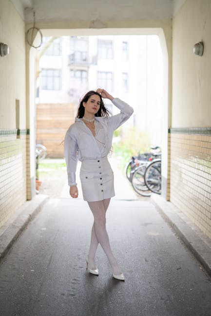 A young woman with long dark hair standing in the middle of a covered passageway or underpass, with beige and yellow tiled walls on either side. She is dressed in a light grey or white buttoned-up blouse with a slightly ruffled collar and a matching skirt, paired with sheer white tights and white high heels. The woman has one hand resting on her head, and her expression appears neutral or contemplative. Behind her, the background shows a residential area with a wooden fence, some greenery, and bicycles parked near a building. The lighting is natural, with daylight illuminating the scene, and the overall atmosphere is calm and urban. The passageway has a curved ceiling with two small wall-mounted lights, and the setting could be relevant for situations involving private waste disposal or waste management, such as rubbish removal services, though no rubbish or waste elements are visible in this image.