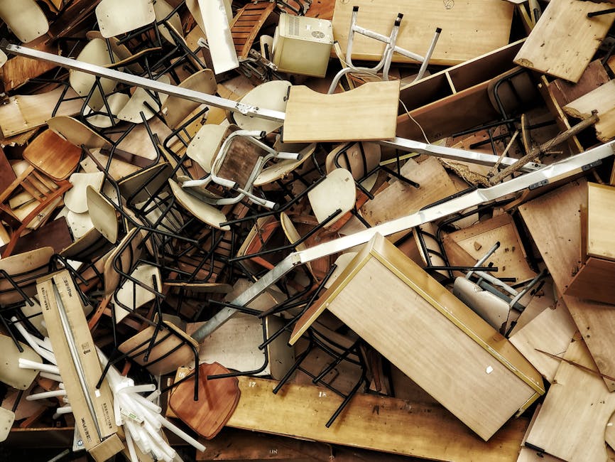 A large collection of broken and discarded furniture and furnishings, including wooden desks with light-colored surfaces, metal-framed chairs with beige and wooden seats and backs, and various small tables. The furniture is haphazardly piled together in a cluttered indoor setting, with some items partially obscured by others. The surfaces show signs of wear, such as scratches, dents, and darkened areas, indicating they are used or damaged. Among the objects, some chairs are stacking or leaning against each other, while desks and tables are placed at odd angles, creating a chaotic scene. The background features additional furniture and discarded items, all densely packed, contributing to the sense of debris accumulation. The environment appears to be a warehouse or storage area ready for disposal or collection by an independent rubbish removal service, reflecting an on-site clearance scenario. The lighting is ambient, evenly illuminating the scene without harsh shadows, emphasizing the array of materials, textures, and the disorganized state of the waste items, relevant to private rubbish disposal and clearance processes by companies like rubbishclearancecollierswood.org.uk.
