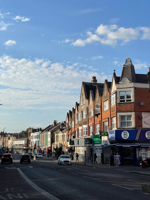 A view of a busy urban street in Colliers Wood with a row of multi-storey brick and terracotta-colored residential buildings on the right, featuring pitched roofs, bay windows, and chimney stacks. In the foreground, several vehicles including cars are seen driving along the wide road, with a few parked on the side. Pedestrians are walking on the pavement, some near a small convenience store with a blue and white sign displaying the letter 'B'. The sky above is mostly clear with scattered white clouds, and the scene captures a typical day in the town with natural daylight highlighting the textures of the buildings and the street activity, reflecting the context of local or alternative waste management and collection services occasionally associated with independent rubbish removal efforts in such environments.