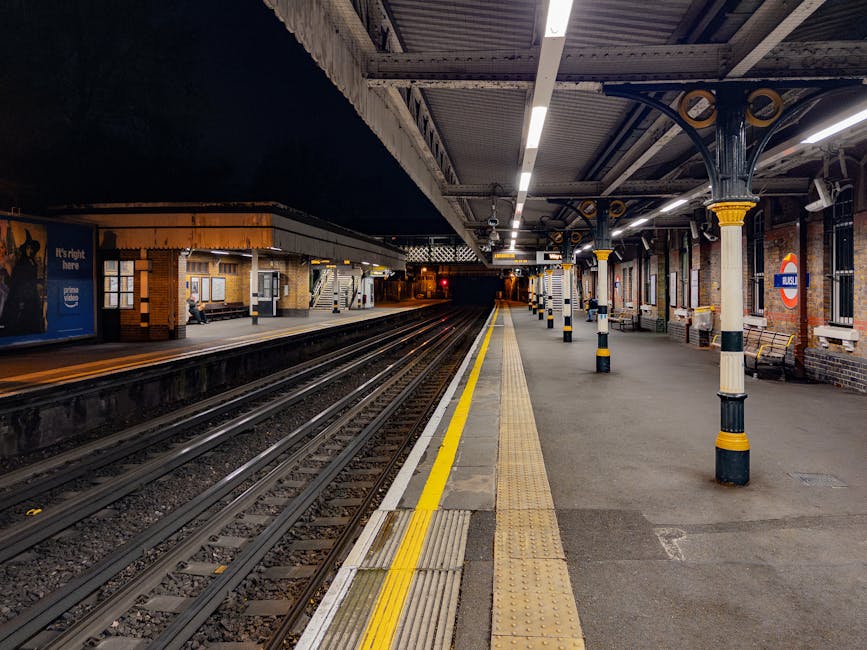 An empty railway station platform at night, illuminated by overhead lighting. The platform features a tactile yellow safety strip along the edge, with textured paving designed for accessibility. Multiple black and cream bollards with yellow stripes are positioned along the covered shelter, which is supported by ornate, dark-colored cast-iron columns with decorative gold accents. The platform's brick wall on the right displays a large round station sign with the red and blue London Underground roundel, indicating the station's name. Wooden benches are placed at intervals under the shelter, and several windows are visible in the station building structure on the left side of the image. The train tracks run parallel to the platform into a dark tunnel or tunnel entrance in the background. The overall scene is quiet with no visible passengers or train activity, capturing a calm, nocturnal atmosphere typical of an urban railway station, often associated with independent or private transport options as part of alternative travel arrangements.