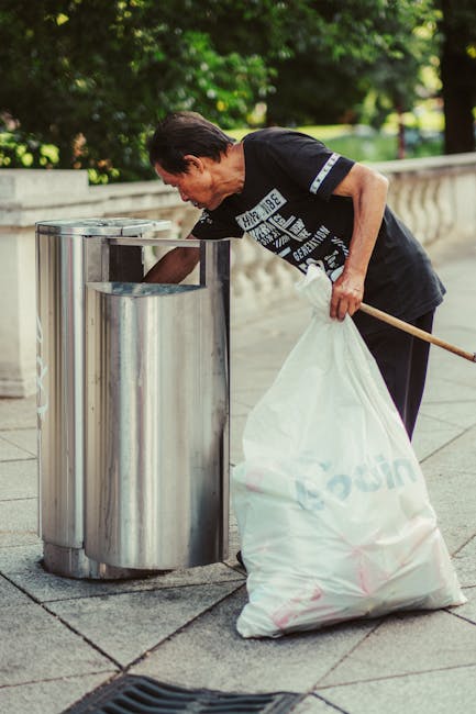 A middle-aged woman with short dark hair, dressed in a black T-shirt with printed white text, is seen litter picking near a set of three cylindrical stainless steel trash bins on a paved sidewalk. She is holding a white plastic bag filled with collected rubbish in her left hand, while her right hand is reaching into the bin to dispose of waste. The scene is set outdoors, with a backdrop of lush green trees and foliage, and a stone wall bordering the walkway. The pavement appears clean and well-maintained, and the woman's posture indicates active participation in waste management or litter control, reflecting a focus on environmental cleanliness. The stainless steel bins are positioned close together, and the overall setting suggests a public or communal outdoor space where independent rubbish collection and disposal are taking place, possibly as part of a community clean-up or scheduled rubbish clearance service from Rubbish Clearance Colliers Wood.