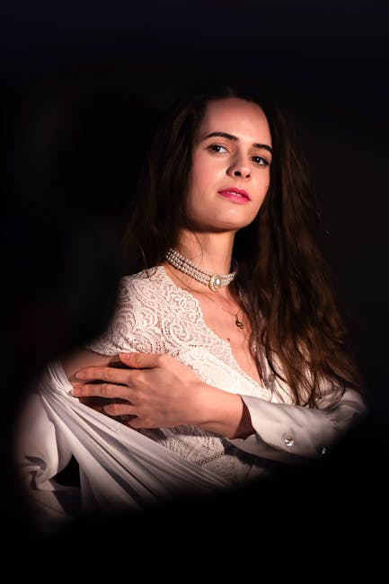 A professional portrait of a woman with long, wavy brown hair and fair skin, set against a dark background. She is wearing a lace-patterned, short-sleeved, white dress with a high neckline, complemented by a multi-strand pearl necklace and a small gold pendant. Her expression is serious, with a slight tilt of her head, and she looks directly at the camera. Her left arm is crossed over her chest, holding the edge of her dress, revealing a smooth, white fabric that drapes over her shoulder. The lighting highlights her facial features and the textures of her clothing, creating a subtle contrast with the dark setting. The overall composition emphasizes her elegant appearance, suitable for a formal or professional context, though there are no elements indicating a specific location or activity related to rubbish removal or waste management.